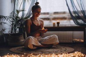 A young woman sitting with her legs crossed in her dimly lit room as she holds a candle and closes her eyes in mediation.